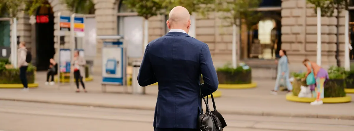 A man in a navy suit walking through a city square, seen from behind